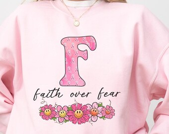 Peut inclure: Sweat-shirt rose pâle avec une lettre F rose et blanche, entourée de fleurs roses avec des visages souriants jaunes. Le texte "faith over fear" est imprimé sous la lettre.
