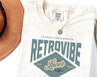 May include: Ivory t-shirt with a blue and white graphic print that says "Classic Fashionista Retrovibe Lover Since 1984".