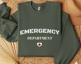 May include: Military green sweatshirt with the words "EMERGENCY DEPARTMENT" in white block letters. A white heart with a red cross is below the text. The sweatshirt is folded on a woven surface.