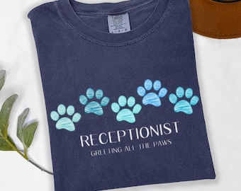 Może przedstawiać: Granatowa koszulka z rzędem niebieskich odcisków łap i napisem "RECEPTIONIST GREETING ALL THE PAWS". Koszulka jest złożona i ma metkę Comfort Colors.