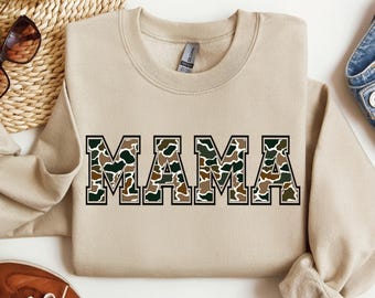 Könnte beinhalten: Ein beiger Pullover mit dem Wort "MAMA" in schwarzen umrandeten Buchstaben bedruckt. Die Buchstaben sind mit einem Tarnmuster in Braun- und Grüntönen gefüllt. Der Pullover liegt auf einer weißen Oberfläche.