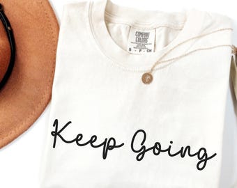 May include: Ivory-colored t-shirt with the words "Keep Going" in black script. A gold necklace with a pendant rests on the shirt. A brown hat is partially visible in the upper left corner.