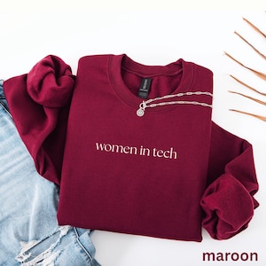 Könnte beinhalten: Ein weinrotes Sweatshirt mit dem weißen Text "women in tech". Eine silberne Halskette mit einem kreisförmigen Anhänger liegt über dem Sweatshirt. Das Wort "maroon" befindet sich unten rechts.