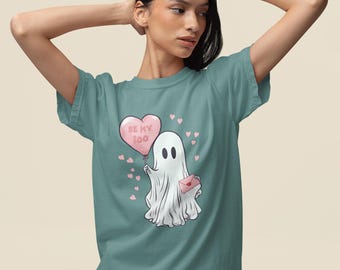 May include: Light green t-shirt featuring a cartoon ghost holding a pink heart-shaped balloon with the text "BE MY BOO". The ghost also holds a pink envelope. Small pink hearts are scattered around the design.