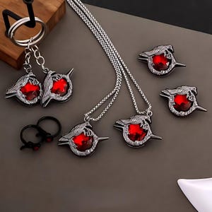 Puede incluir: Un conjunto de joyas con un colgante de plata con una piedra preciosa roja y un diseño de cuervo. El colgante está disponible como collar, llavero y anillo.