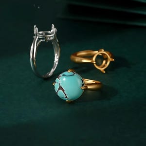 以下が含まれることがあります： ターコイズの宝石をセットした、銀と金のリング3つ。リングは緑色の表面の上に置かれています。