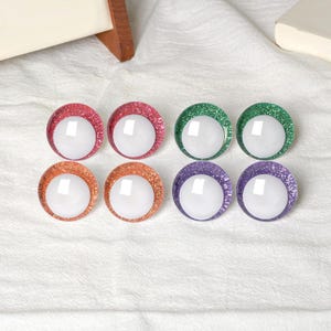 May include: Eight colorful, round craft eyes with glitter rims in red, green, orange, and purple. Each eye has a white, reflective center. The eyes are arranged in two rows of four on a white fabric surface.