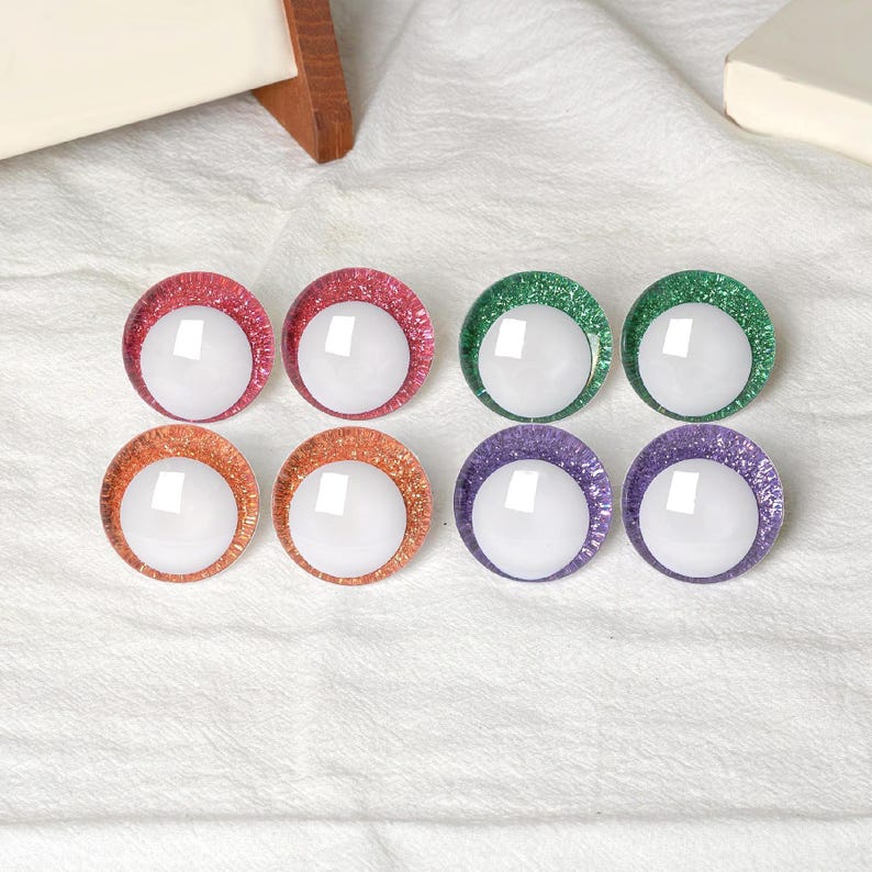 May include: Eight colorful, round craft eyes with glitter rims in red, green, orange, and purple. Each eye has a white, reflective center. The eyes are arranged in two rows of four on a white fabric surface.