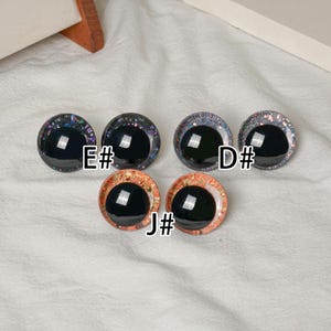 Pu&ograve; includere: Sei occhi di bambola rotondi e lucidi, neri, con bordi decorativi. I bordi presentano glitter e accenti colorati. Gli occhi sono etichettati con le lettere "E#", "D#" e "J#".