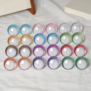 May include: A collection of colorful, glitter-rimmed, round craft embellishments. The assortment includes various colors such as blue, pink, silver, gold, brown, purple, red, orange, and green. Each embellishment has a white, circular center.