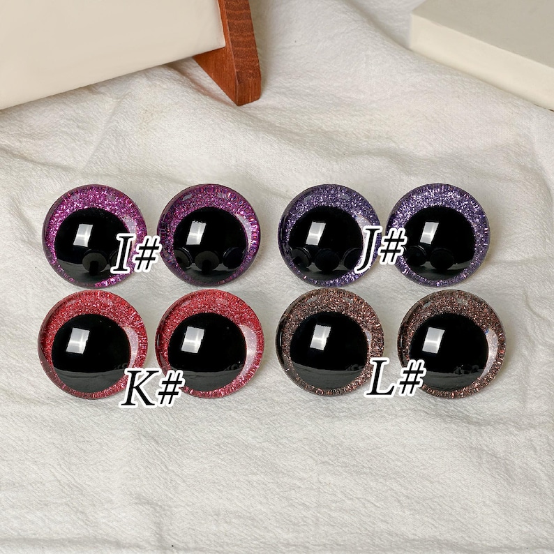 May include: Eight round craft eyes with black pupils and glittery irises in pink, purple, red, and brown. The pairs are labeled I#, J#, K#, and L#. Each eye features a smooth, glossy finish and a distinct sparkling texture around the center.