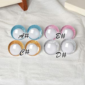 May include: A collection of four pairs of round, glitter-rimmed doll eyes in various colors: blue, pink, gold, and silver. Each eye has a white, reflective center. The letters A#, B#, C#, and D# are printed on the image.
