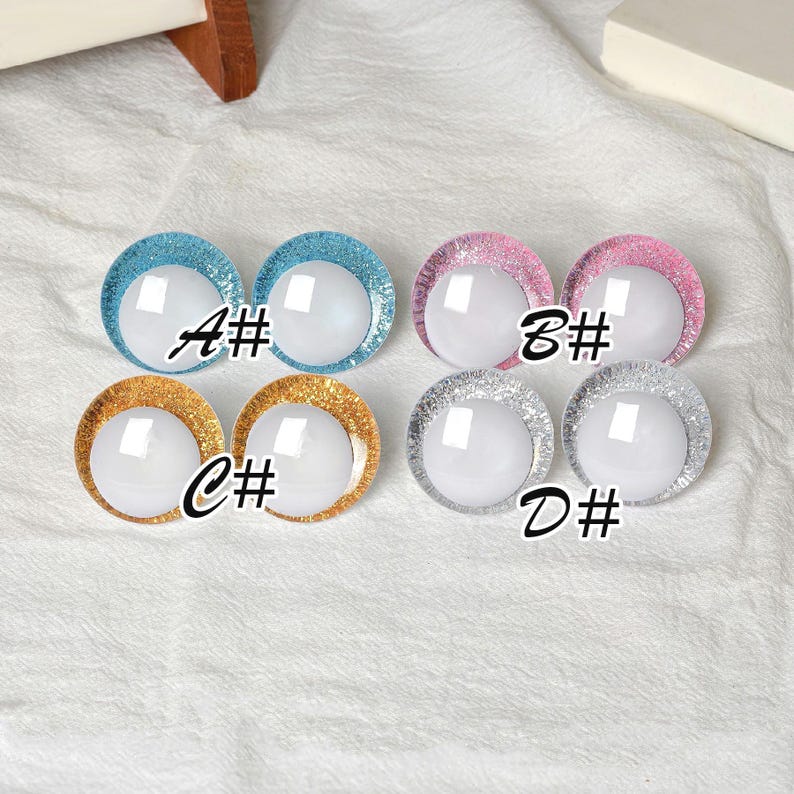 May include: A collection of four pairs of round, glitter-rimmed doll eyes in various colors: blue, pink, gold, and silver. Each eye has a white, reflective center. The letters A#, B#, C#, and D# are printed on the image.