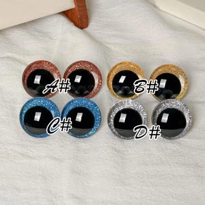 May include: Eight circular craft eyes with black pupils and glittery iris rings in four color pairs: copper labeled A#, gold labeled B#, blue labeled C#, and silver labeled D#. Each eye features a smooth, reflective surface and a distinct glitter texture.