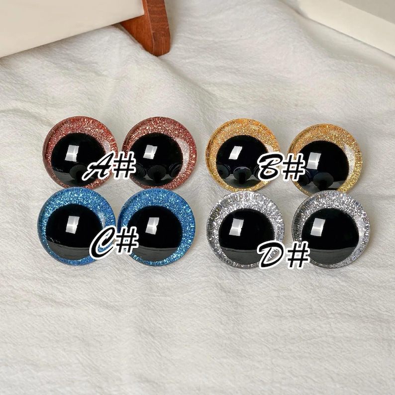 May include: Eight circular craft eyes with black pupils and glittery iris rings in four color pairs: copper labeled A#, gold labeled B#, blue labeled C#, and silver labeled D#. Each eye features a smooth, reflective surface and a distinct glitter texture.
