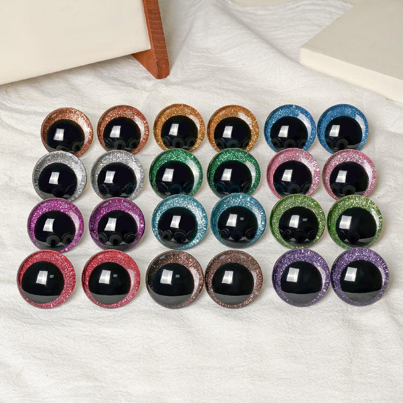 May include: A collection of twenty-four circular craft eyes arranged in four rows of six. Each eye features a large, glossy black pupil surrounded by a vibrant, glittery iris in various colors including red, blue, green, purple, and gold.