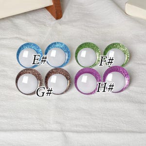 May include: A collection of eight round, glittery glass eyes in various colors: blue, green, brown, and purple. Each eye has a white center and a letter and number designation: E#, F#, G#, and H#.