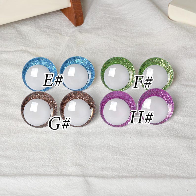 May include: A collection of eight round, glittery glass eyes in various colors: blue, green, brown, and purple. Each eye has a white center and a letter and number designation: E#, F#, G#, and H#.