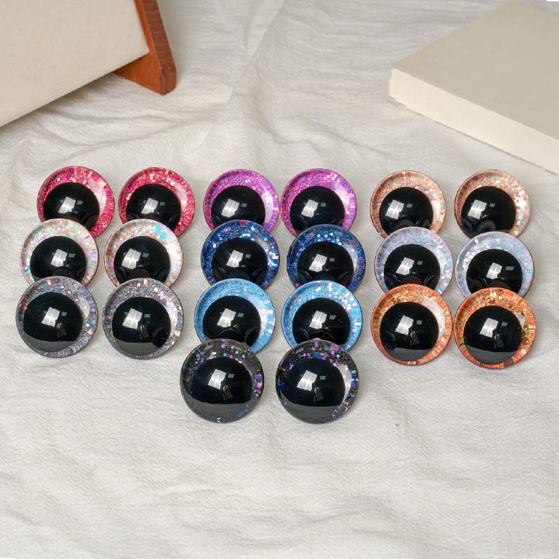 Pu&ograve; includere: Una collezione di occhi di vetro colorati con pupille nere e bordi glitterati. Gli occhi sono in vari colori, tra cui rosa, viola, blu e arancione. Sono disposti su una superficie bianca, adatti per lavori artigianali e creazione di bambole.