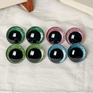 May include: Eight circular craft eyes with black pupils and glittery irises in shades of green, pink, and blue. Each eye features a smooth, reflective surface with a distinct white light reflection, arranged in two rows of four on a white fabric.