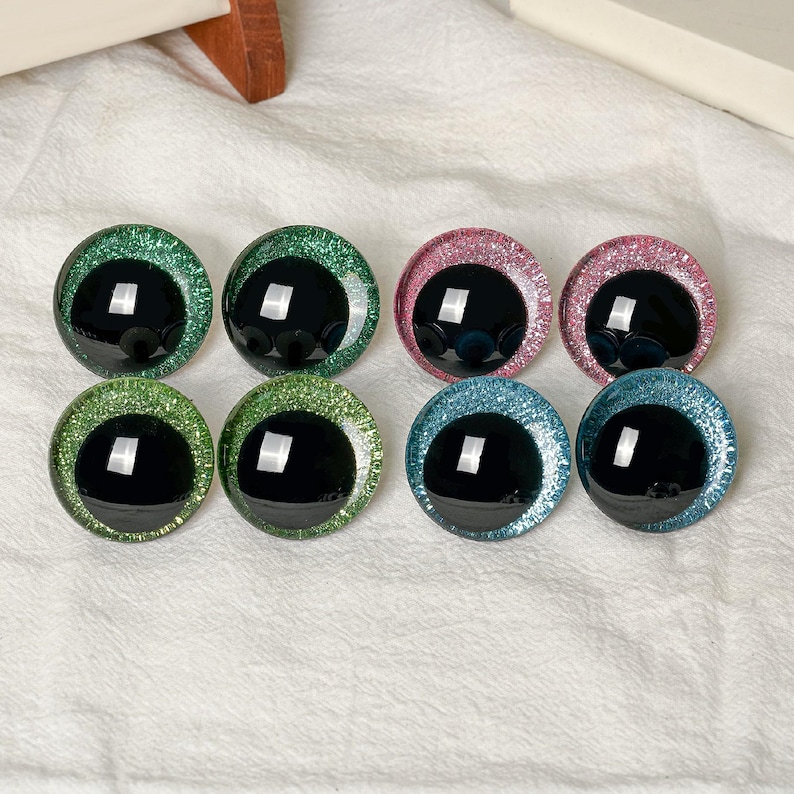 May include: Eight circular craft eyes with black pupils and glittery irises in shades of green, pink, and blue. Each eye features a smooth, reflective surface with a distinct white light reflection, arranged in two rows of four on a white fabric.
