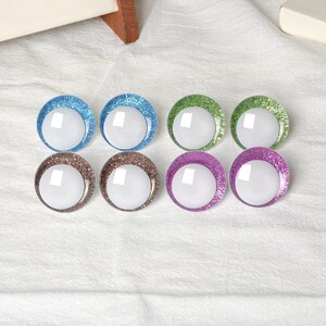 May include: Eight colorful, round craft eyes with glitter rims in blue, green, brown, and purple. Each eye has a white, reflective center. The eyes are arranged in two rows of four on a white, textured surface.