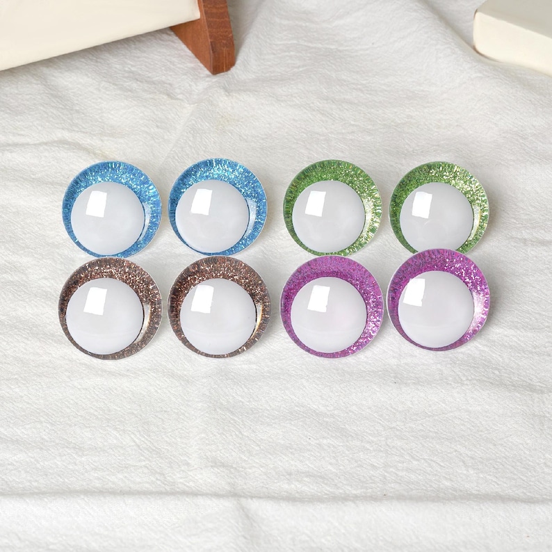 May include: Eight colorful, round craft eyes with glitter rims in blue, green, brown, and purple. Each eye has a white, reflective center. The eyes are arranged in two rows of four on a white, textured surface.