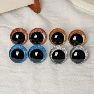 May include: Eight circular craft eyes arranged in two rows. The top row features two copper and two gold glitter eyes. The bottom row includes two blue glitter eyes and two silver glitter eyes. Each eye has a large, smooth, black center pupil.