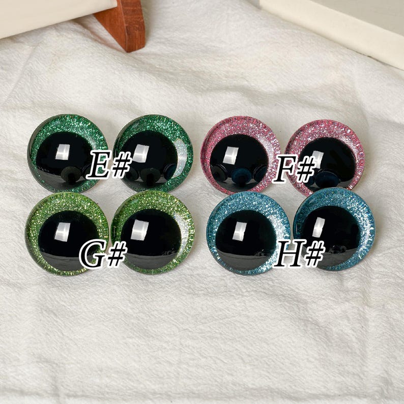 May include: Eight circular craft eyes with black pupils and glittery irises in green, pink, and blue. The pairs are labeled E#, F#, G#, and H#. Each eye features a smooth, domed surface with a reflective finish, arranged in two rows of four.