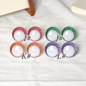 May include: A collection of four pairs of round, glitter-rimmed craft eyes in various colors. The colors include red, green, orange, and purple, each with a white center. Each pair is labeled with a letter and a number.