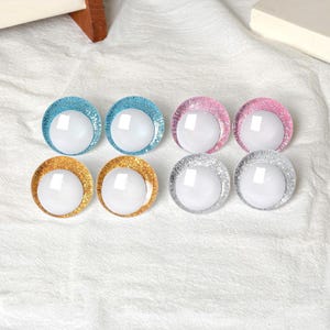 May include: Eight round, decorative craft eyes in various colors. The eyes have a white, spherical center surrounded by a glittery border in blue, pink, gold, and silver. The eyes are arranged on a white, textured surface.