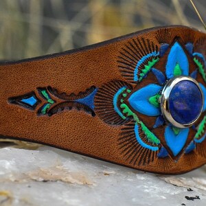 Puede incluir: Una pulsera de cuero marr&oacute;n con una piedra preciosa azul central. La pulsera presenta intrincados dise&ntilde;os florales pintados en azul y verde. La piedra preciosa est&aacute; engastada en un bisel plateado.