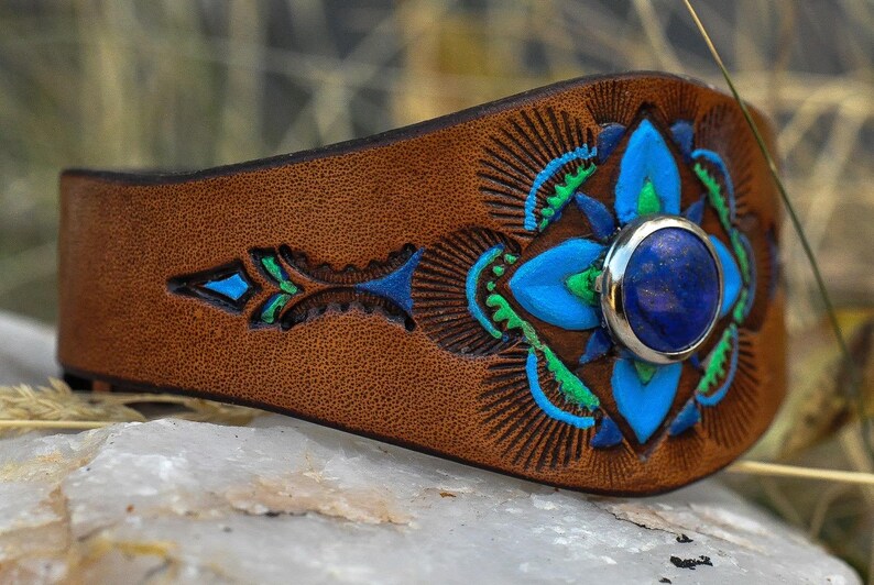 Puede incluir: Una pulsera de cuero marr&oacute;n con una piedra preciosa azul central. La pulsera presenta intrincados dise&ntilde;os florales pintados en azul y verde. La piedra preciosa est&aacute; engastada en un bisel plateado.