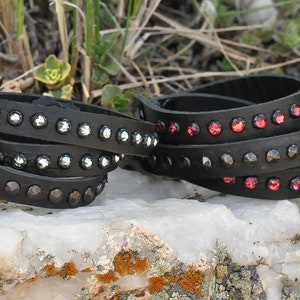 May include: Two black leather bracelets with rows of silver and red rhinestones. The bracelets are wrapped around each other and are lying on a white rock.