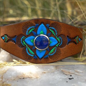 Puede incluir: Una pulsera de cuero marr&oacute;n con un dise&ntilde;o floral azul y verde central y una piedra azul redonda grande. La pulsera tiene un patr&oacute;n tallado detallado, sobre un fondo natural.