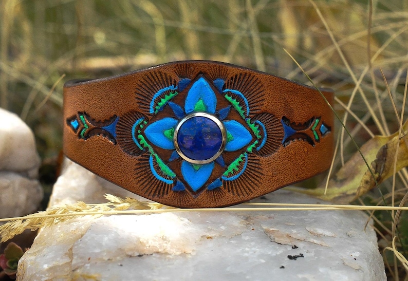 Puede incluir: Una pulsera de cuero marr&oacute;n con un dise&ntilde;o floral azul y verde central y una piedra azul redonda grande. La pulsera tiene un patr&oacute;n tallado detallado, sobre un fondo natural.