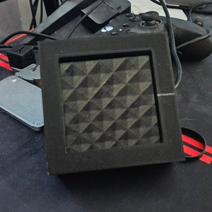 Peut inclure: Un appareil carré noir avec une surface texturée à motifs de losanges. L'appareil est encadré par un bord noir. D'autres éléments en arrière-plan incluent une manette de jeu, une souris et d'autres petits objets gris.