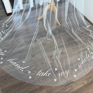 以下が含まれることがあります： 「Take my hand, take my whole life too」という筆記体の文字が書かれた、白いウェディングベール。小さな花の装飾と散りばめられたビーズがベールの縁を飾っています。木製のスタンドに展示されています。