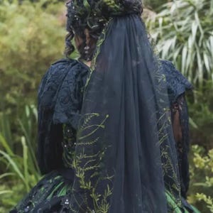 Könnte beinhalten: Ein dunkelgrünes und schwarzes Kleid mit passendem Schleier. Der Schleier ist mit zarten grünen Blumenstickereien verziert. Das Kleid hat komplizierte Spitzendetails an den Ärmeln und am Oberteil, was einen eleganten und ätherischen Look erzeugt. Der Gesamtstil deutet auf ein gotisches oder Fantasy-Thema hin.