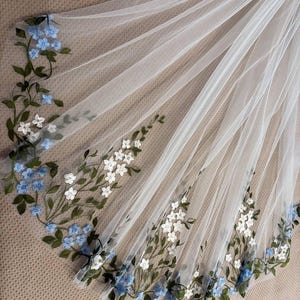 Op de afbeelding: Een witte tule sluier met geborduurde bloemen. De sluier heeft een delicaat patroon van blauwe en witte bloemen met groene bladeren langs de randen. De sluier is gedrapeerd, wat de textuur en het ontwerp laat zien.