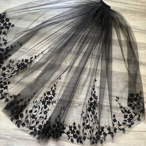 Può includere: Un velo di tulle nero con ricami floreali neri. Il velo è lungo e fluido, con un delicato motivo floreale.