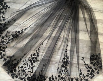 May include: A black tulle veil with black floral embroidery. The veil is long and flowing, with a delicate floral pattern.