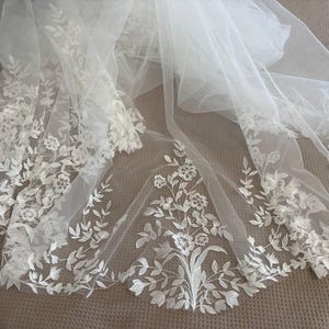 May include: A white wedding veil with intricate floral lace detailing. The delicate lace features a pattern of flowers and leaves, adding a touch of elegance. The veil is made of a sheer, flowing fabric.