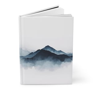 Könnte beinhalten: Ein weißes Notizbuch mit Landschaftsdesign. Das Cover zeigt ein Aquarellgemälde von dunkelblauen Bergen und nebligem Nebel. Das Notizbuch ist geöffnet und zeigt leere weiße Seiten.