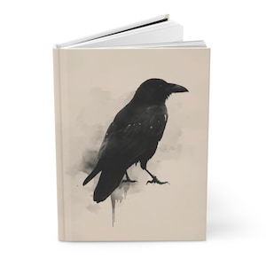 May include: A hardcover journal with a black and white illustration of a raven. The raven is perched on a light beige background with a watercolor effect. The journal is open, revealing blank white pages.