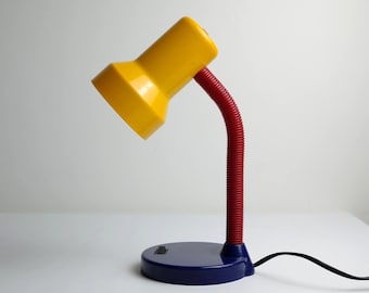 Puede incluir: Una lámpara de escritorio amarilla con cuello flexible rojo y base azul. La lámpara está en ángulo, mostrando su diseño. El cable es negro. La lámpara es de estilo retro.