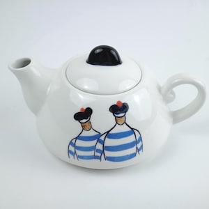 May include: White ceramic teapot with a black knob on the lid and a curved handle. The teapot features a whimsical design of two figures in blue and white striped shirts and black hats.