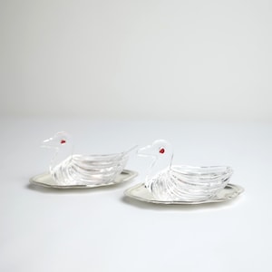 Può includere: Due piatti a forma di anatra in vetro trasparente con accenti rossi sugli occhi, appoggiati su vassoi color argento. Le anatre hanno un design a coste e i vassoi hanno un bordo smerlato. Lo sfondo è bianco.