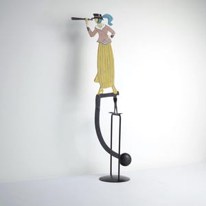 Peut inclure: Une sculpture métallique fantaisiste représentant une figure avec un télescope, portant un chapeau et une longue jupe. La figure est perchée sur une structure métallique noire incurvée avec une base ronde et un petit bougeoir.