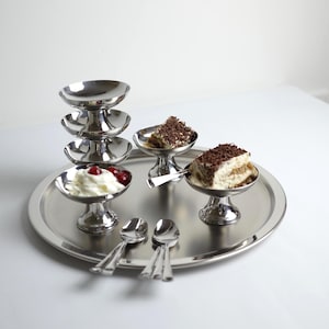 Puede incluir: Una bandeja de acero inoxidable con cuencos de postre y cucharas. Los cuencos contienen postres, como crema batida con cerezas y tiramisú. También hay una pila de cuencos vacíos.